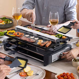 A multi-functional electric raclette grill  in use at a dining table, with people holding a dish and turning the control dial. 