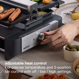 A close-up of a countertop raclette grill  with a hand adjusting the front dial. Grilled sausages and veggies are cooking on the top plates, and bowls of food and a plate are visible around it.
