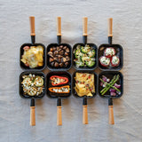 Nine small black frying pans with various food items on a textured surface