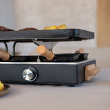 Black raclette grill with wooden handles on a kitchen counter.