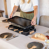 Person setting up a black electric raclette grill on a dining table with plates and utensils.